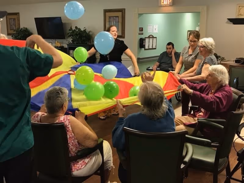 A group of elderly individuals and caregivers sitting in a circle indoors, holding the edges of a colorful parachute with green and blue balloons bouncing on top of it, engaging in a group activity.