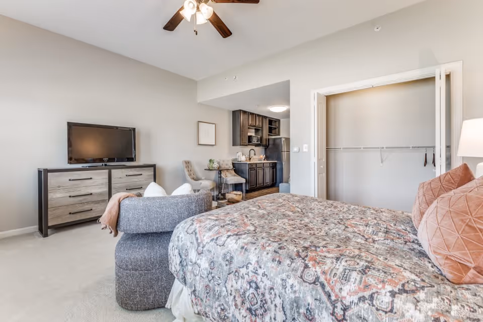 A bright and spacious senior living bedroom featuring a large bed with patterned bedding and multiple pillows. The room includes a gray upholstered chair with a pink throw, a dresser with a flat-screen TV on top, and an open closet with white bi-fold doors. In the background, there is a small kitchenette with dark wood cabinets, a microwave, and a refrigerator, along with a small dining area with two chairs and a round table. The ceiling has a fan with lights, and the walls are painted a light neutral color.