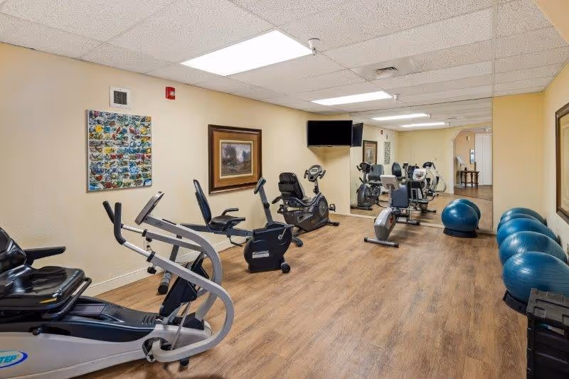 A senior living community fitness room with exercise equipment including stationary bikes, elliptical machines, and several blue exercise balls lined up against the wall. The room has wood-style flooring, beige walls with framed artwork, a large mirror on one side, and a wall-mounted TV.