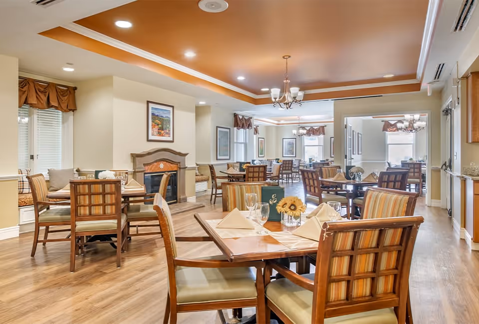 Well-appointed dining room with multiple wooden tables and chairs, place settings, chandeliers, and warm ceiling accents.