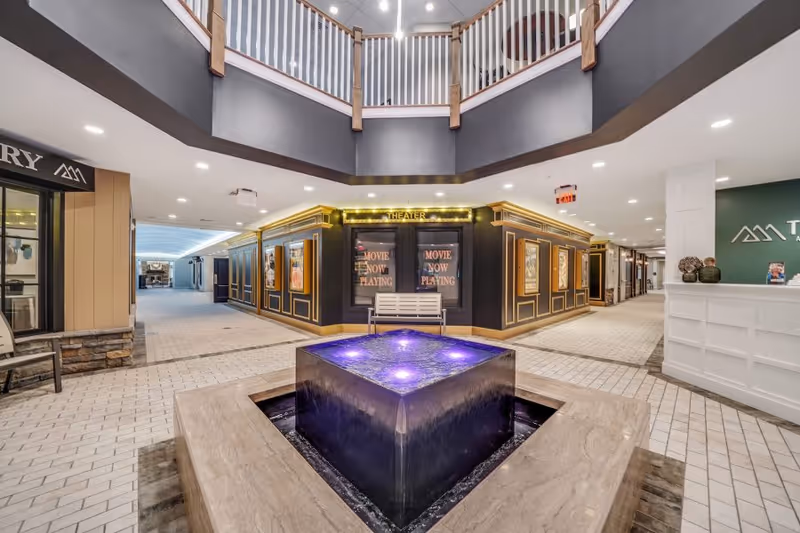 Atrium-style interior lobby with a central illuminated square fountain, a theater entrance and reception desk in the background.