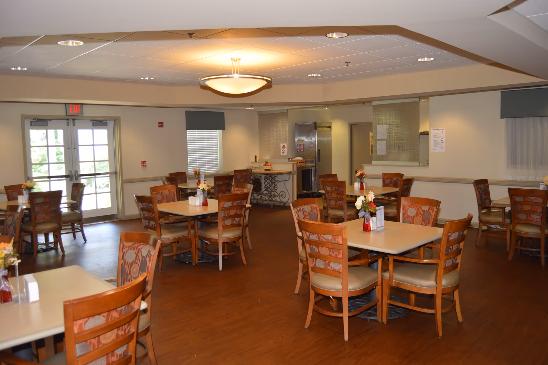 A dining room with multiple square tables and wooden chairs with patterned cushions. Each table has a small flower arrangement and a menu or information card. The room has wood flooring, beige walls, and a ceiling with recessed lighting and a central light fixture. There are windows with blinds and a double door exit with glass panels.