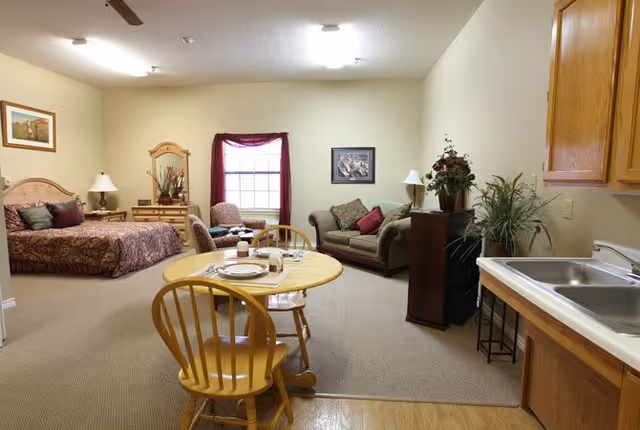 A spacious senior living facility room featuring a bed with a wooden headboard and bedside table with a lamp, a small round dining table with two wooden chairs set with plates and cups, a cozy sitting area with a loveseat and armchair, a window with burgundy curtains, and a kitchenette with a sink and wooden cabinets.