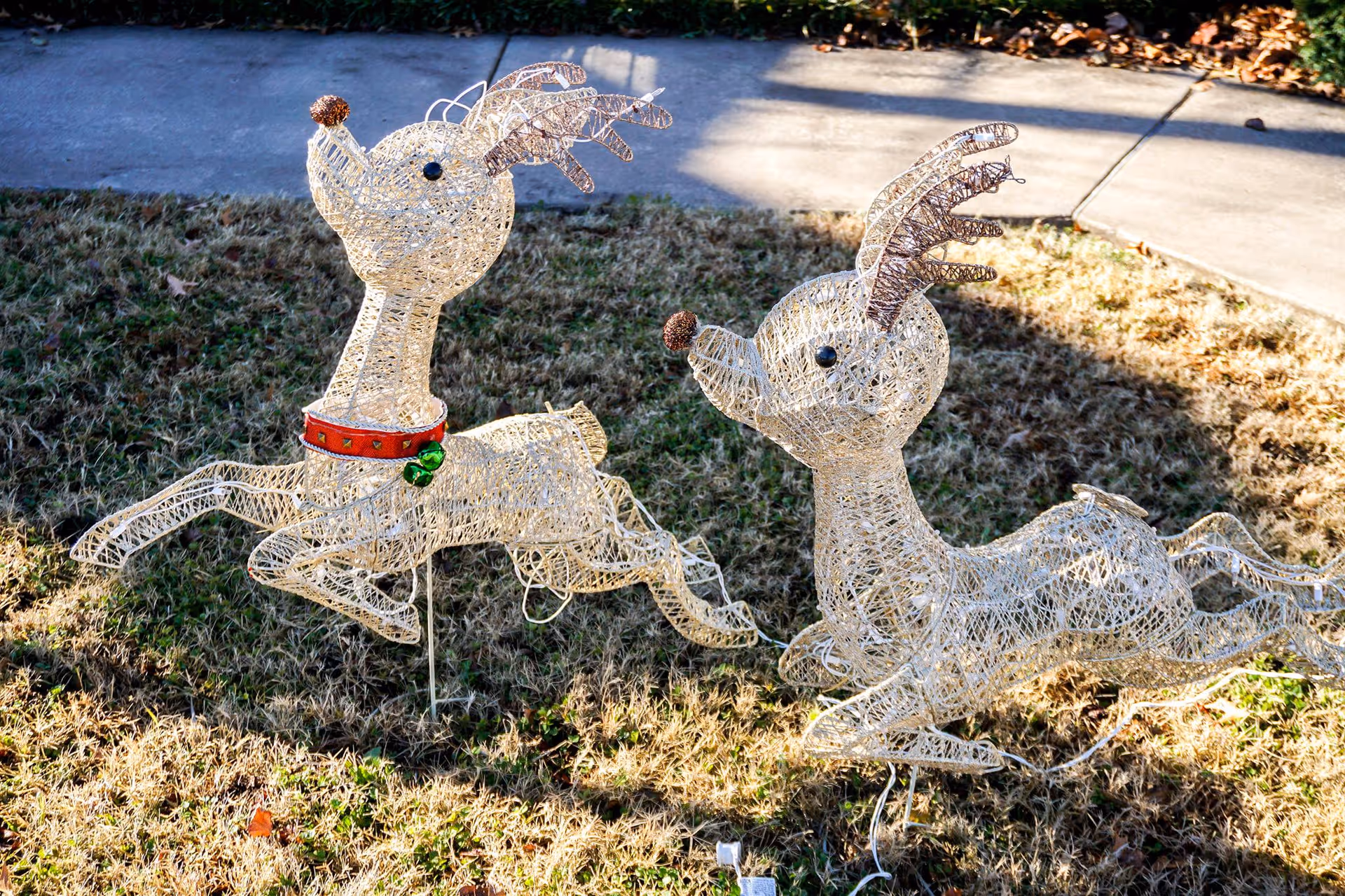 Two decorative wireframe reindeer figures placed on grass near a sidewalk, one with a red collar and green bells, both appearing to be in a leaping pose.