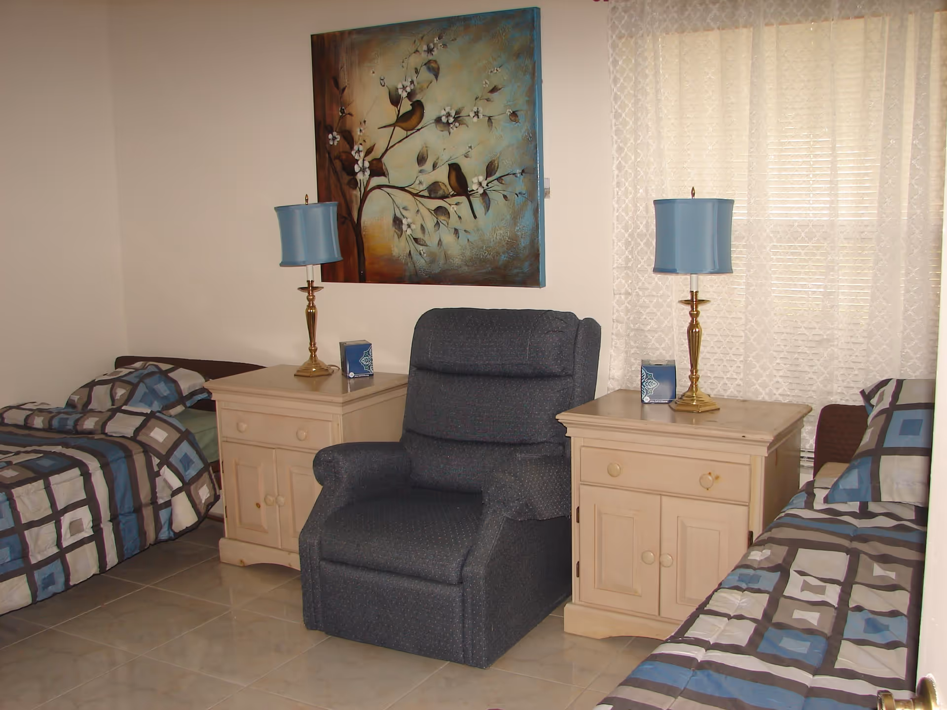 A bedroom with two single beds on either side of a dark blue armchair. Each bed has a geometric patterned bedspread in shades of blue, gray, and white. Between each bed and the armchair is a light wooden nightstand with a blue lampshade on a brass lamp and a box of tissues. A large painting of birds on a branch hangs on the wall above the armchair, and a window with sheer white curtains is visible behind the right nightstand.