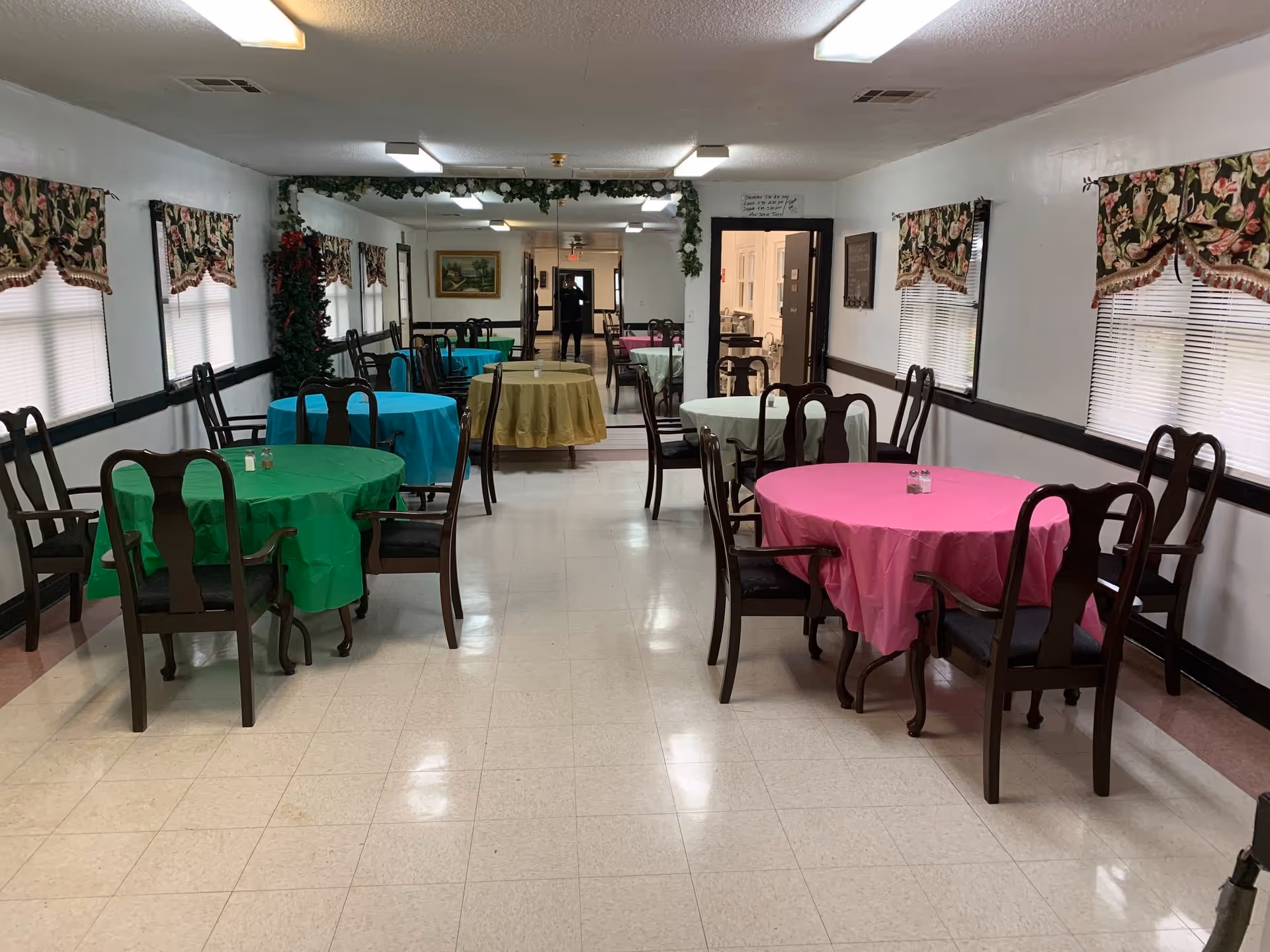 A dining room with several round tables covered in colorful tablecloths in green, blue, yellow, and pink. Each table is surrounded by wooden chairs with armrests. The room has white walls with floral valances on the windows and a large mirror on the far wall reflecting the room. The floor is tiled, and there are fluorescent lights on the ceiling.