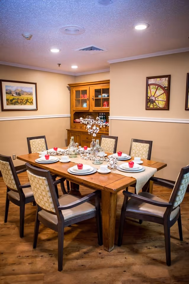 A dining room with a wooden table set for six people. Each place setting has a white plate, cup, and a red apple on the plate. The table is decorated with glass bottles and a cotton branch centerpiece. There are six chairs with patterned upholstery around the table. In the background, there is a wooden cabinet with glass doors and framed pictures on the beige walls.