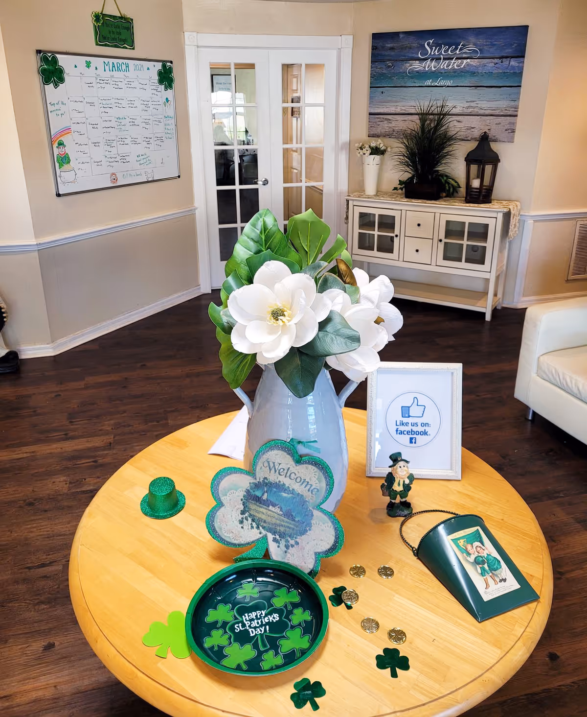 Interior view of a senior living facility common area decorated for St. Patrick's Day with a round wooden table in the foreground displaying white flowers in a vase, a green glittery shamrock welcome sign, a small green hat, a plate with shamrocks and 'Happy St. Patrick's Day!' text, gold coins, and a small leprechaun figurine. In the background, there is a white cabinet with plants and a lantern on top, a framed sign encouraging to like on Facebook, a wall calendar for March 2021, and a large wall art with the text 'Sweet Water at Largo'.