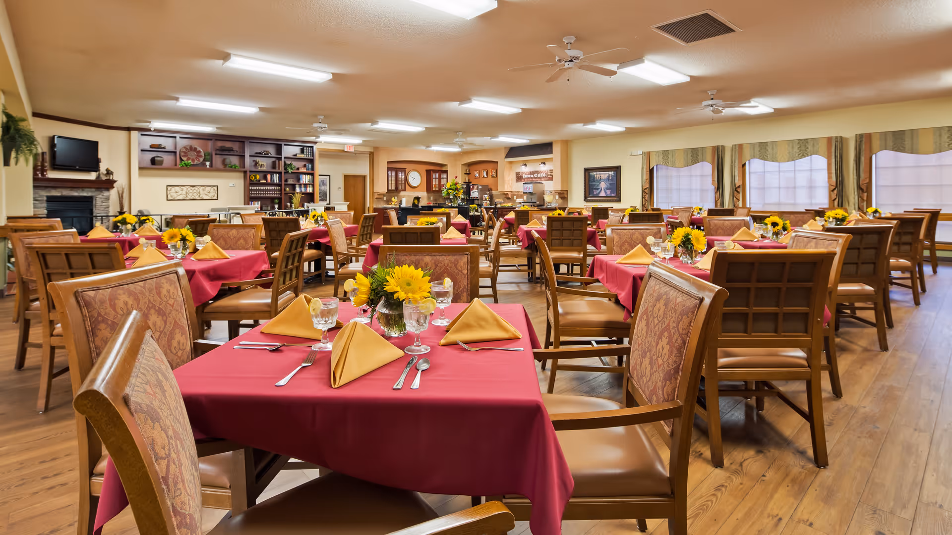 A spacious dining room with multiple tables covered in red tablecloths and set with yellow napkins, silverware, and glasses of water with lemon. Each table has a centerpiece of sunflowers. The room has wooden floors, ceiling fans, and large windows with curtains. In the background, there is a coffee station labeled 'Java Café' and a fireplace with a mounted TV above it.