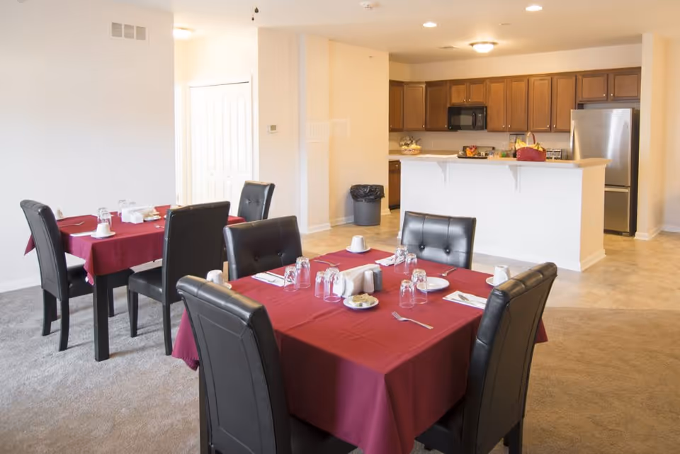 Dining area with two tables covered in red tablecloths, each set with plates, cups, utensils, and glasses. The room has black cushioned chairs around the tables. In the background, there is a kitchen with wooden cabinets, a stainless steel refrigerator, a microwave, and a countertop with some items on it.