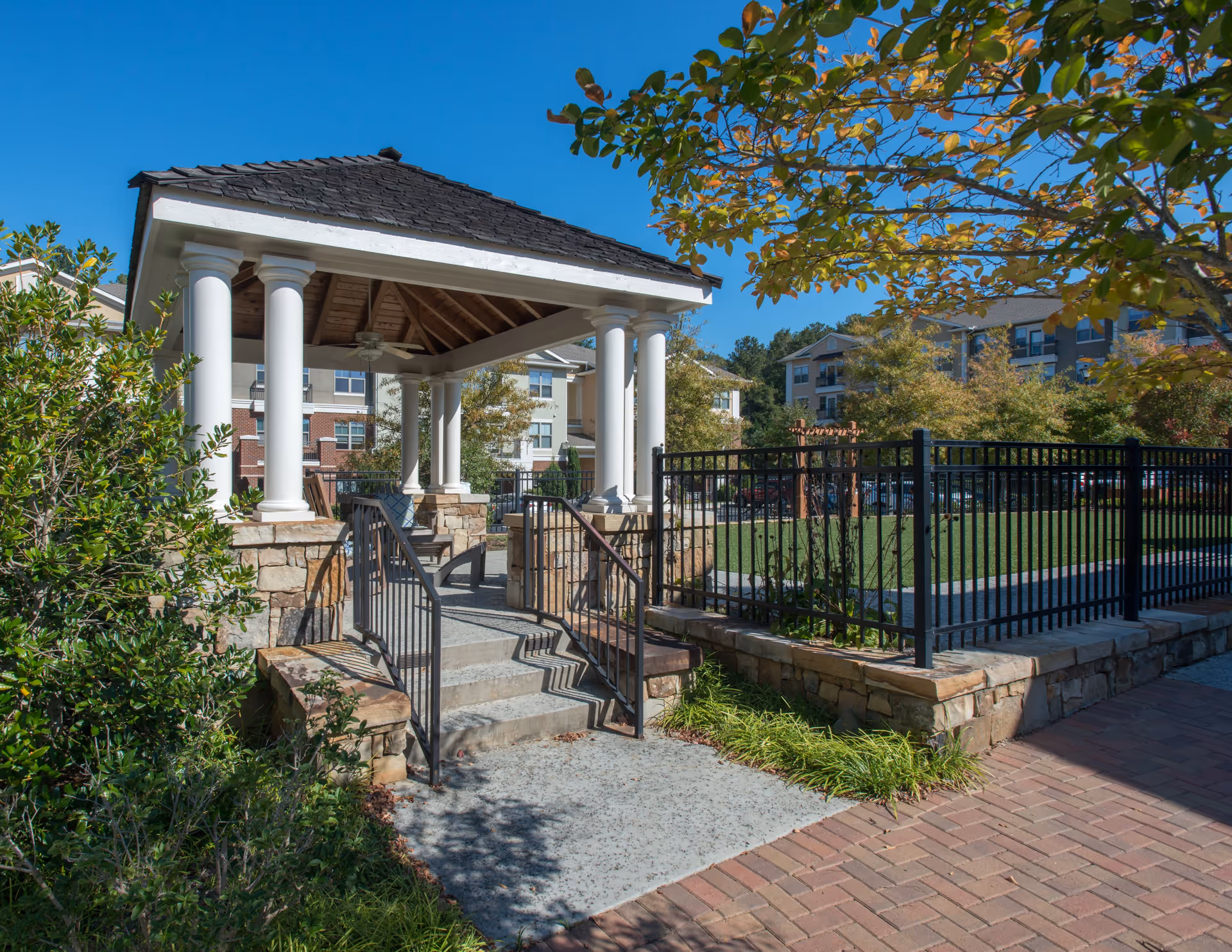 Outdoor gazebo with white columns and stone base, surrounded by greenery and a black metal fence, with residential buildings in the background under a clear blue sky.