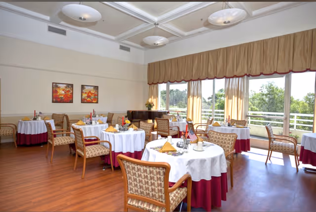 Bright dining room with round tables set with white tablecloths and napkins, upholstered chairs, and large windows overlooking greenery.