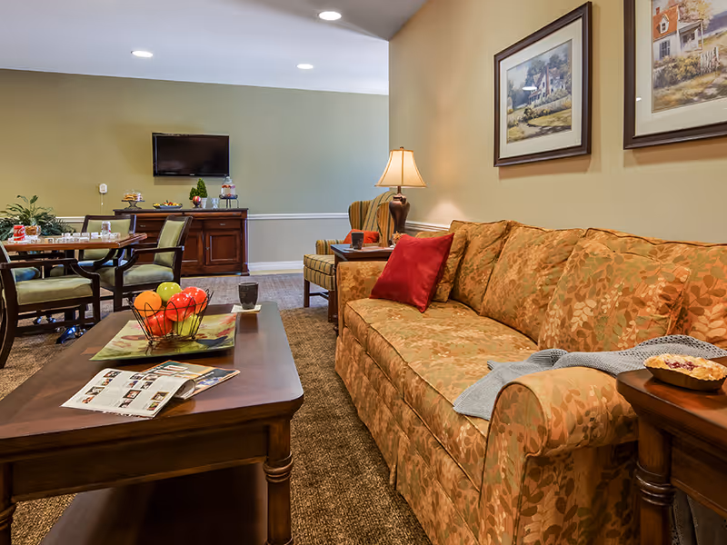 A cozy living room area in an assisted living facility featuring a patterned sofa with red and orange cushions, a wooden coffee table with a fruit basket and magazines, a side table with a lamp and a pie, a dining table with chairs in the background, a wall-mounted TV, and framed artwork on the walls.