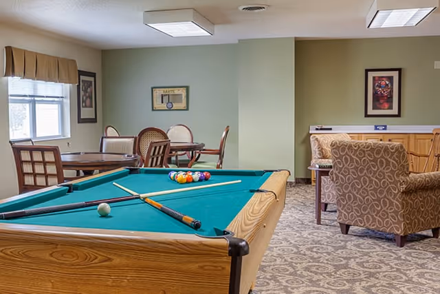 Recreation room with a pool table in the foreground, seating and dining tables and framed art on green walls.