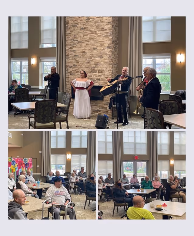 A mariachi band performs in a spacious room with large windows and a stone fireplace. The band includes a woman in a white dress and three men playing violin, guitar, and trumpet. Several elderly residents sit at tables watching the performance, some using walkers and wearing masks.