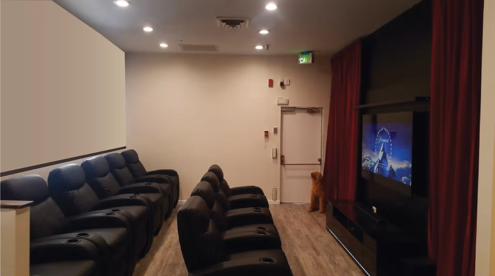 A small theater room with two rows of black leather recliner chairs facing a large screen on the wall. The screen is framed by red curtains, and a door is visible at the back of the room. The floor is wooden, and the ceiling has recessed lighting.