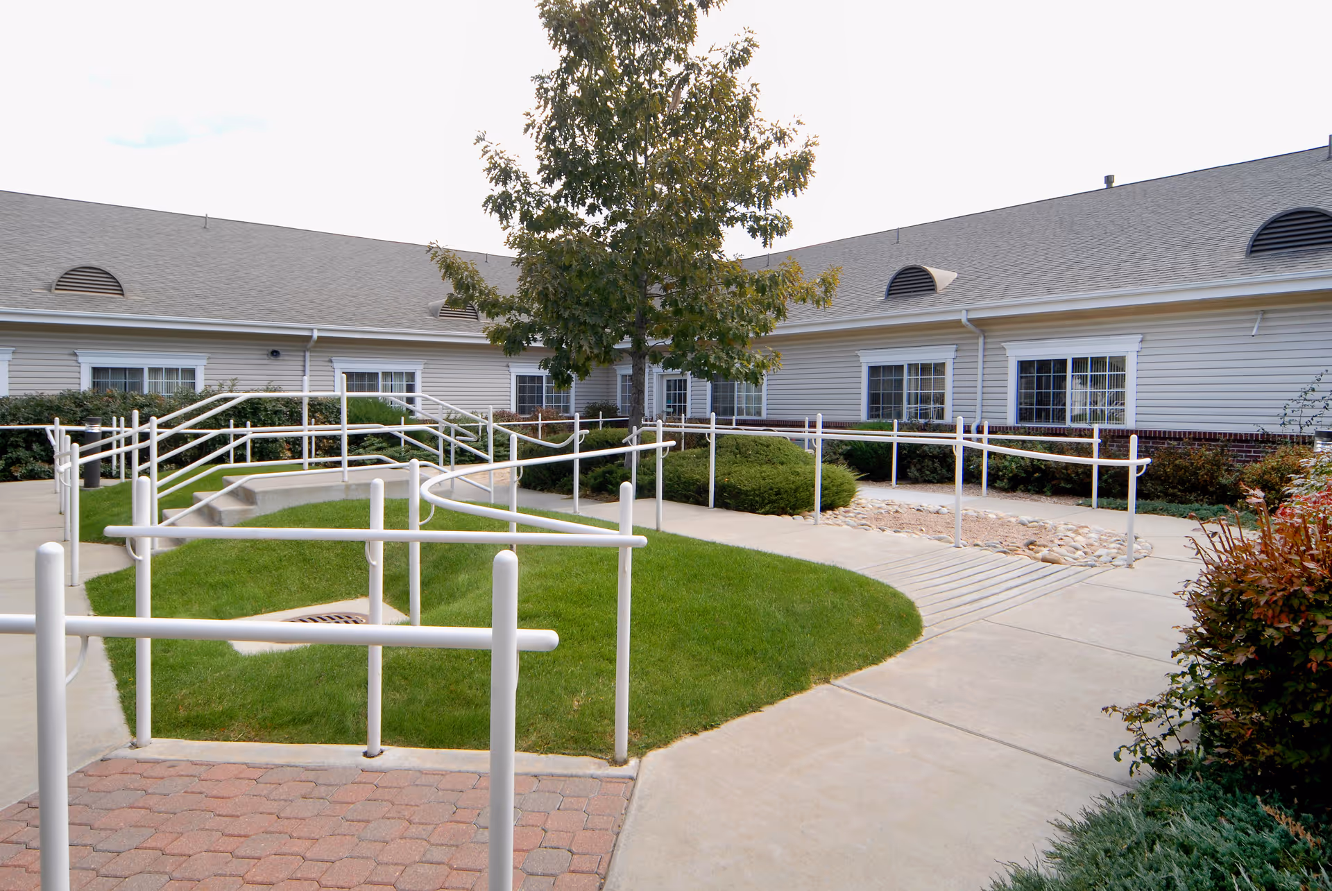 Outdoor courtyard with paved walkways, white handrail ramps, grassy islands and a tree in front of a single-story building.