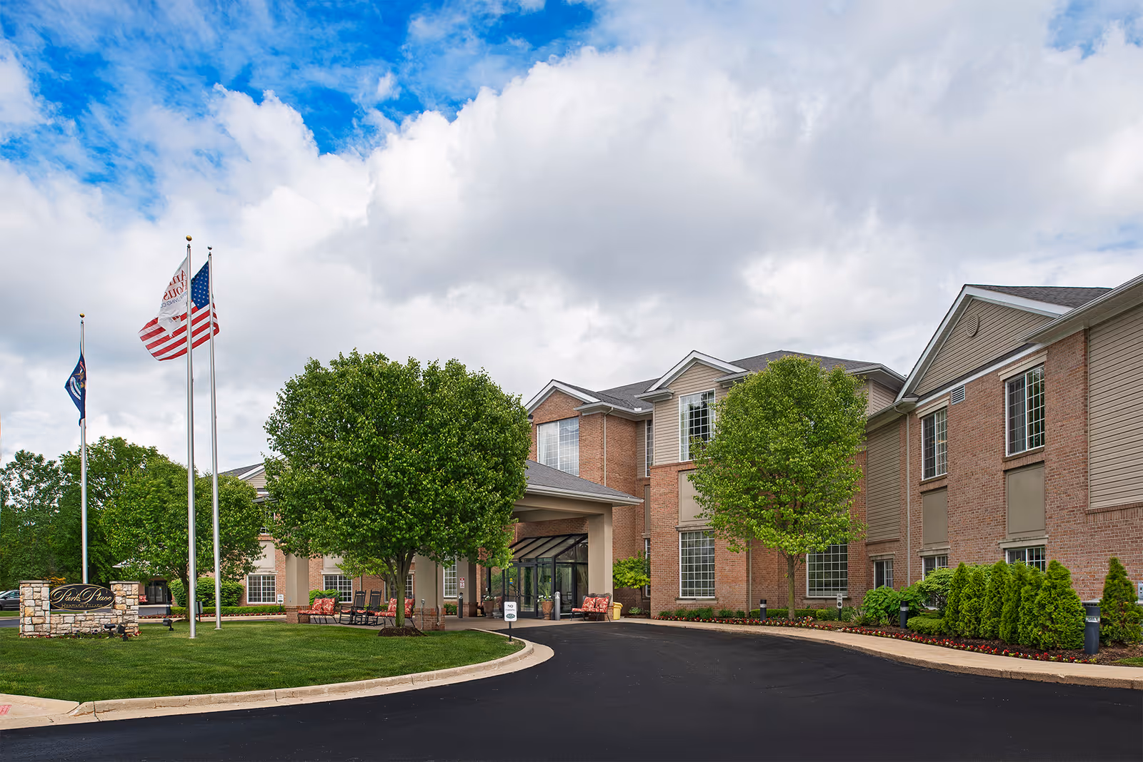 Exterior view of a senior living facility named American House Park Place, showing a two-story brick building with large windows, a covered entrance, three flagpoles with flags, green trees, and a well-maintained lawn under a partly cloudy sky.