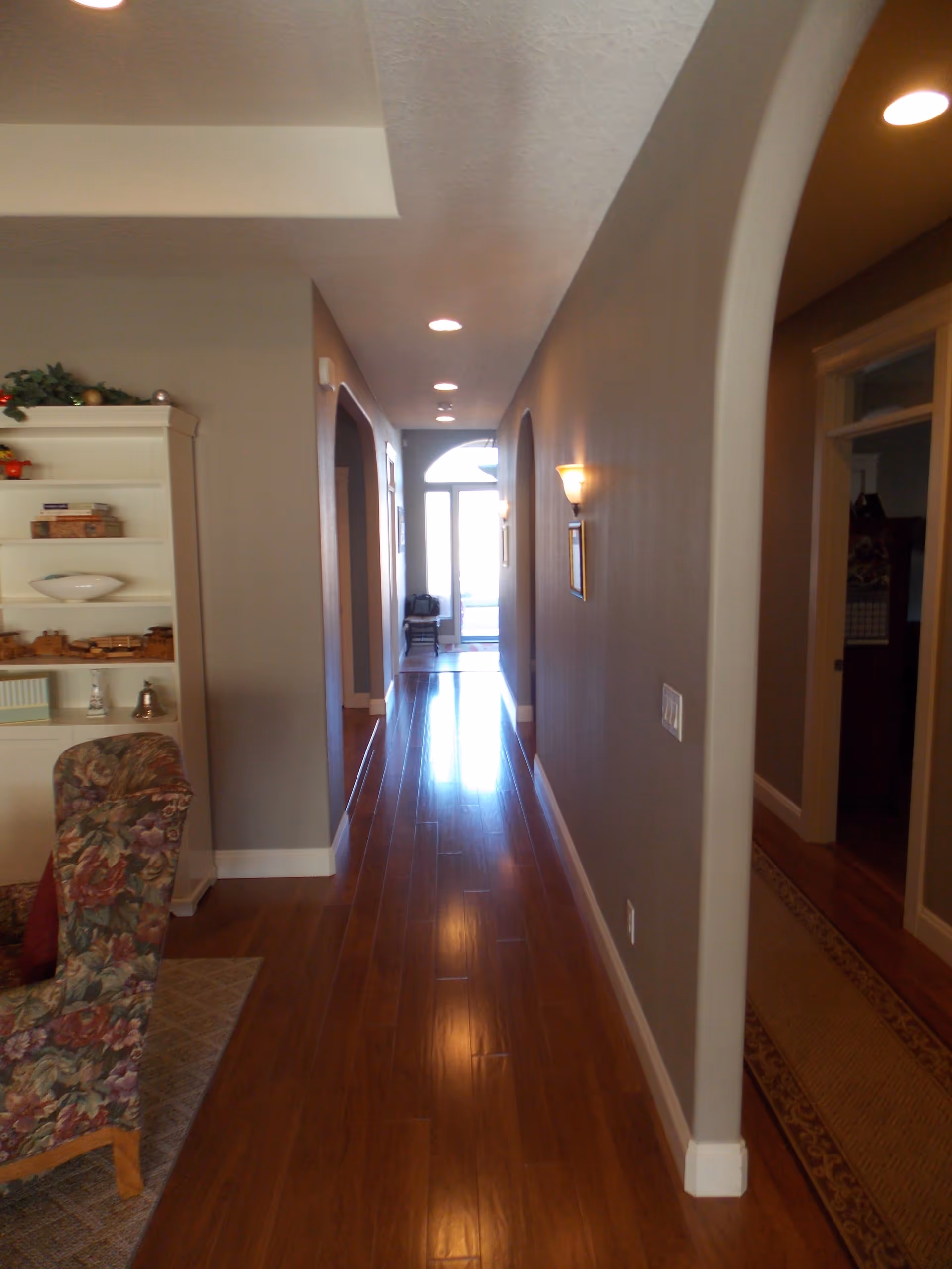 A long, narrow hallway with wooden flooring and gray walls, illuminated by recessed ceiling lights and wall sconces. On the left side, there is a floral upholstered chair and a white shelving unit with decorative items. The hallway leads to a door with a large window letting in natural light.
