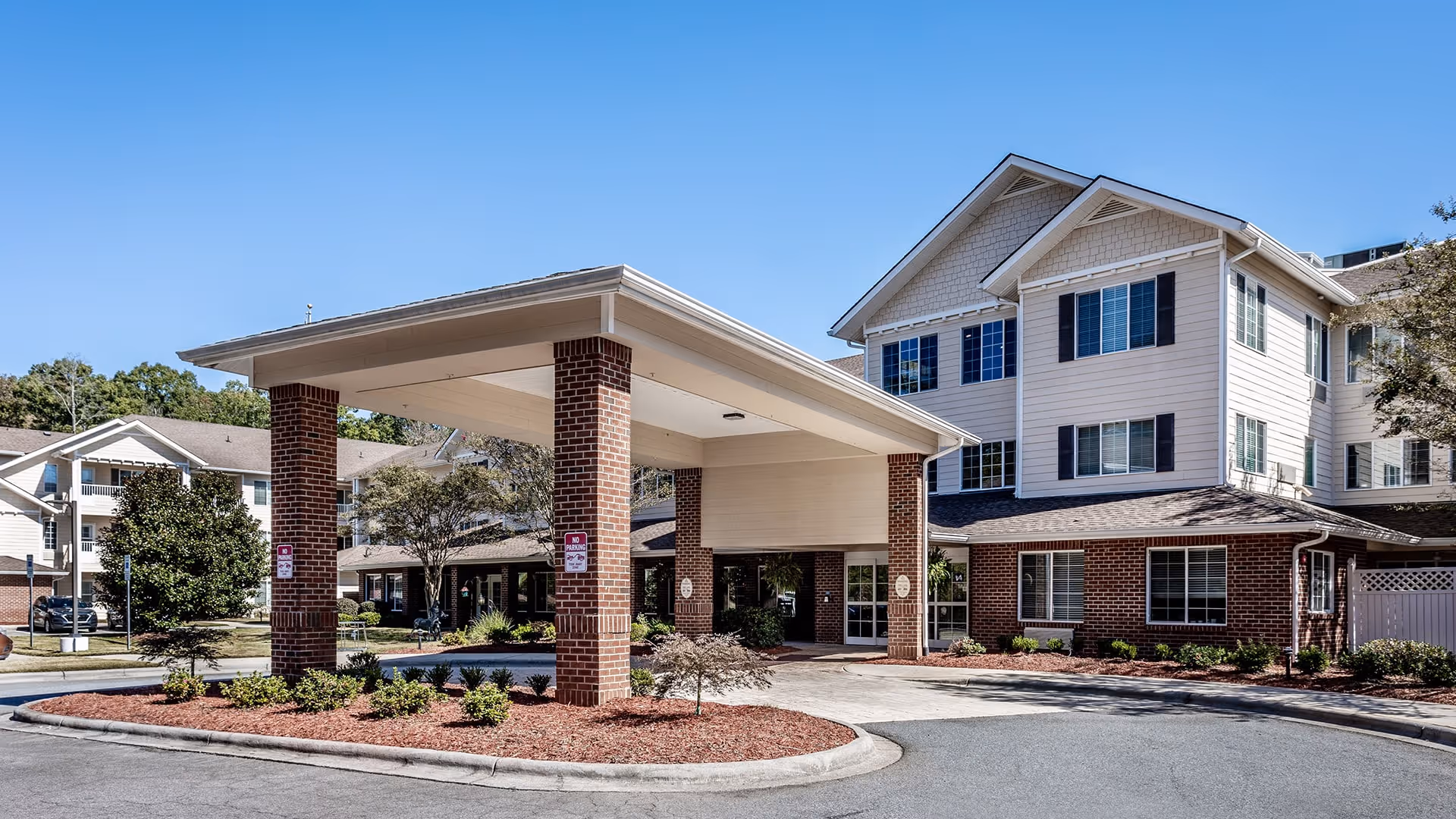 Exterior view of a senior living facility named Holiday Willow Grove, showing a covered entrance supported by brick pillars, a driveway, landscaped areas with small bushes and trees, and a multi-story building with white siding and black shutters under a clear blue sky.