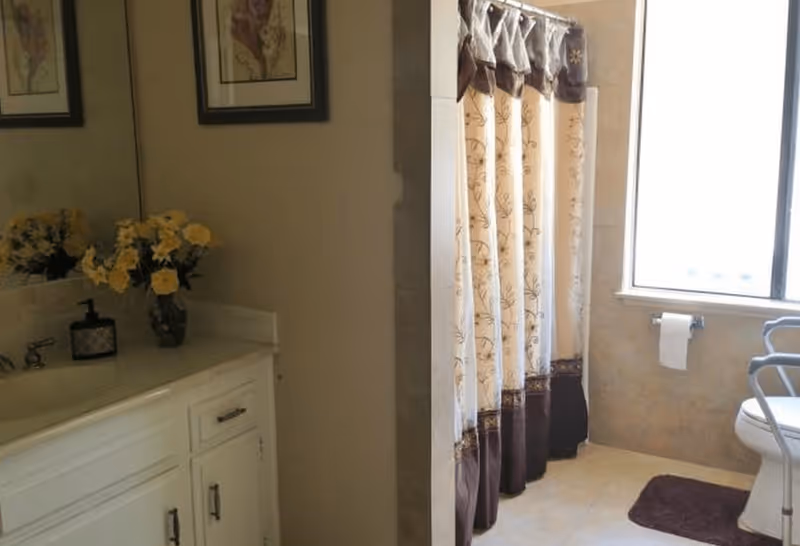 Bathroom with a white vanity cabinet topped with a vase of yellow flowers and a soap dispenser. There is a shower area with a decorative curtain, a window with a toilet paper holder beneath it, and a toilet with a safety frame around it. A brown bath mat is placed on the tiled floor.