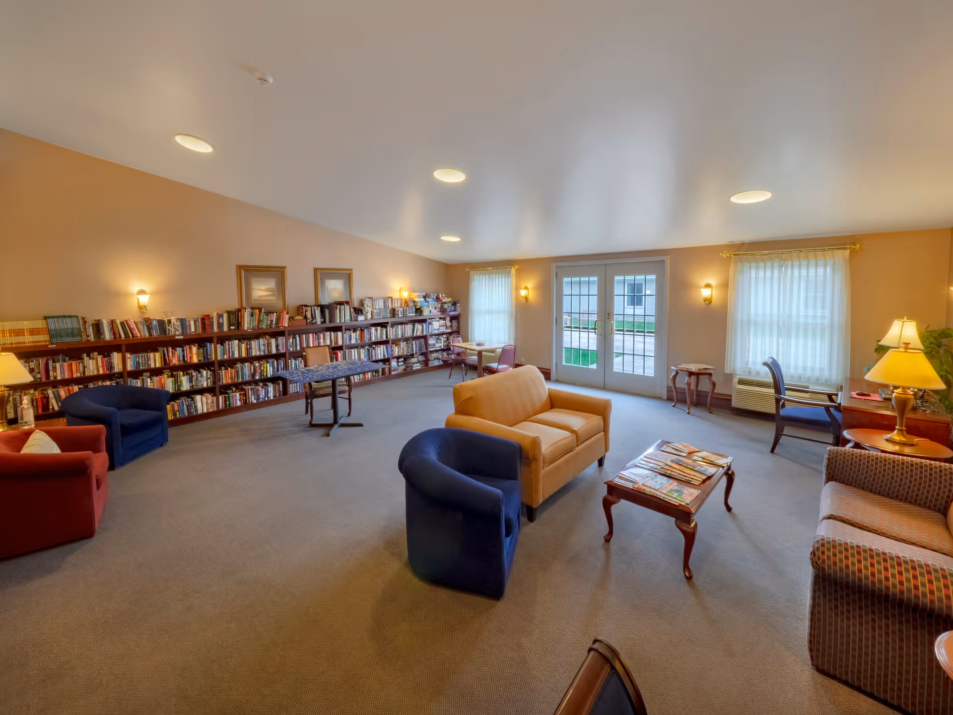 A spacious and well-lit common area with multiple seating options including armchairs and sofas arranged around a coffee table with magazines. The room features large windows with sheer curtains, a set of double glass doors leading outside, and a long bookshelf filled with books along one wall. There are also small tables and chairs near the windows and warm lighting fixtures on the walls.