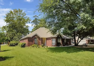 A single-story brick building with a beige roof surrounded by green grass and trees under a blue sky with some clouds.