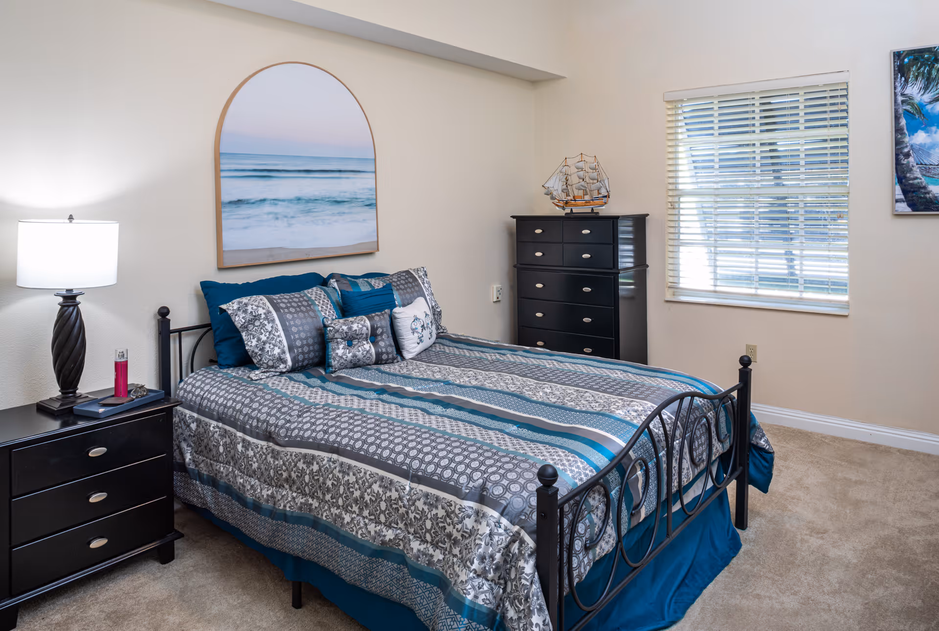 A neatly made bed with a patterned blue, gray, and white bedspread and multiple pillows in a bedroom. To the left of the bed is a black nightstand with a lamp and some small items. On the wall above the bed is a large framed picture of a calm ocean scene. To the right of the bed is a black dresser with a model ship on top. A window with white blinds is on the right wall, letting in natural light. The room has beige walls and carpeted floor.
