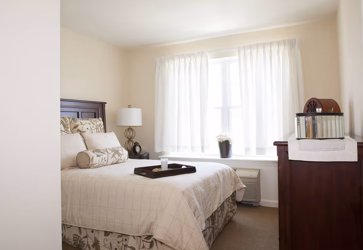 A bright and cozy bedroom with a neatly made bed featuring beige and floral patterned bedding. There is a dark wooden headboard, a bedside table with a lamp and an alarm clock, and a window with white curtains letting in natural light. A dark wooden dresser with a decorative item on top is positioned near the window.