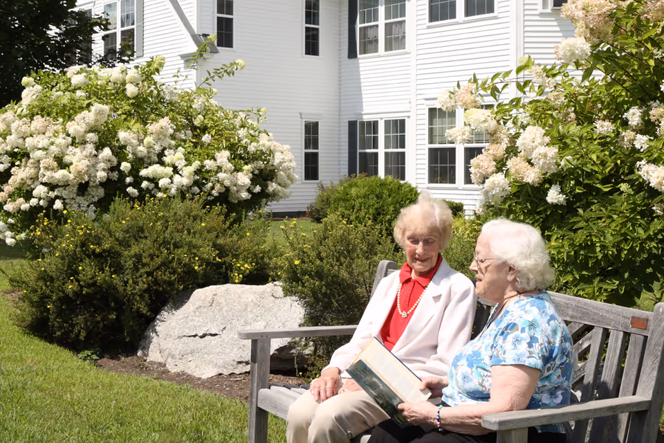 Two elderly women sitting on a wooden bench outside near flowering bushes and greenery, with a white building with multiple windows in the background. One woman is holding an open book.