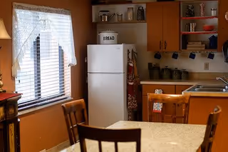 Cozy kitchen area with wooden cabinets, a white refrigerator labeled 'BREAD', a sink, and a dining table with wooden chairs. A window with blinds and a lace curtain lets in natural light.