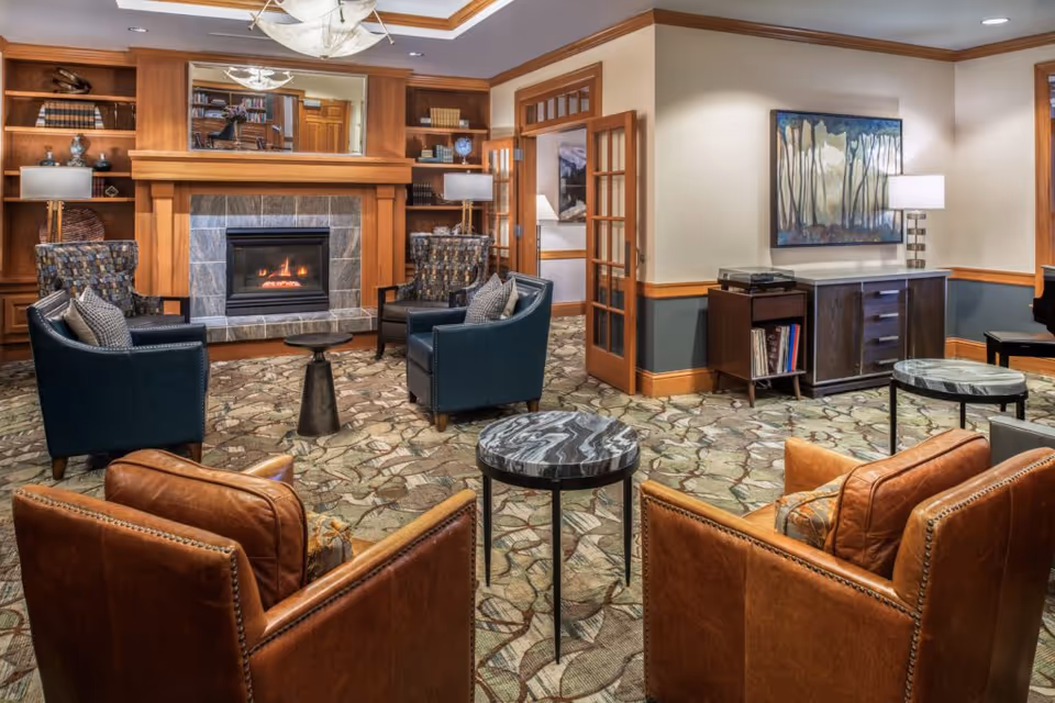 A cozy living room area with a lit fireplace surrounded by wooden built-in shelves filled with books and decorative items. The room features a patterned carpet, four leather and upholstered armchairs arranged around two small round tables with marble tops. There is a wooden cabinet with a lamp and artwork on the wall, and a doorway with glass-paneled double doors leading to another room.