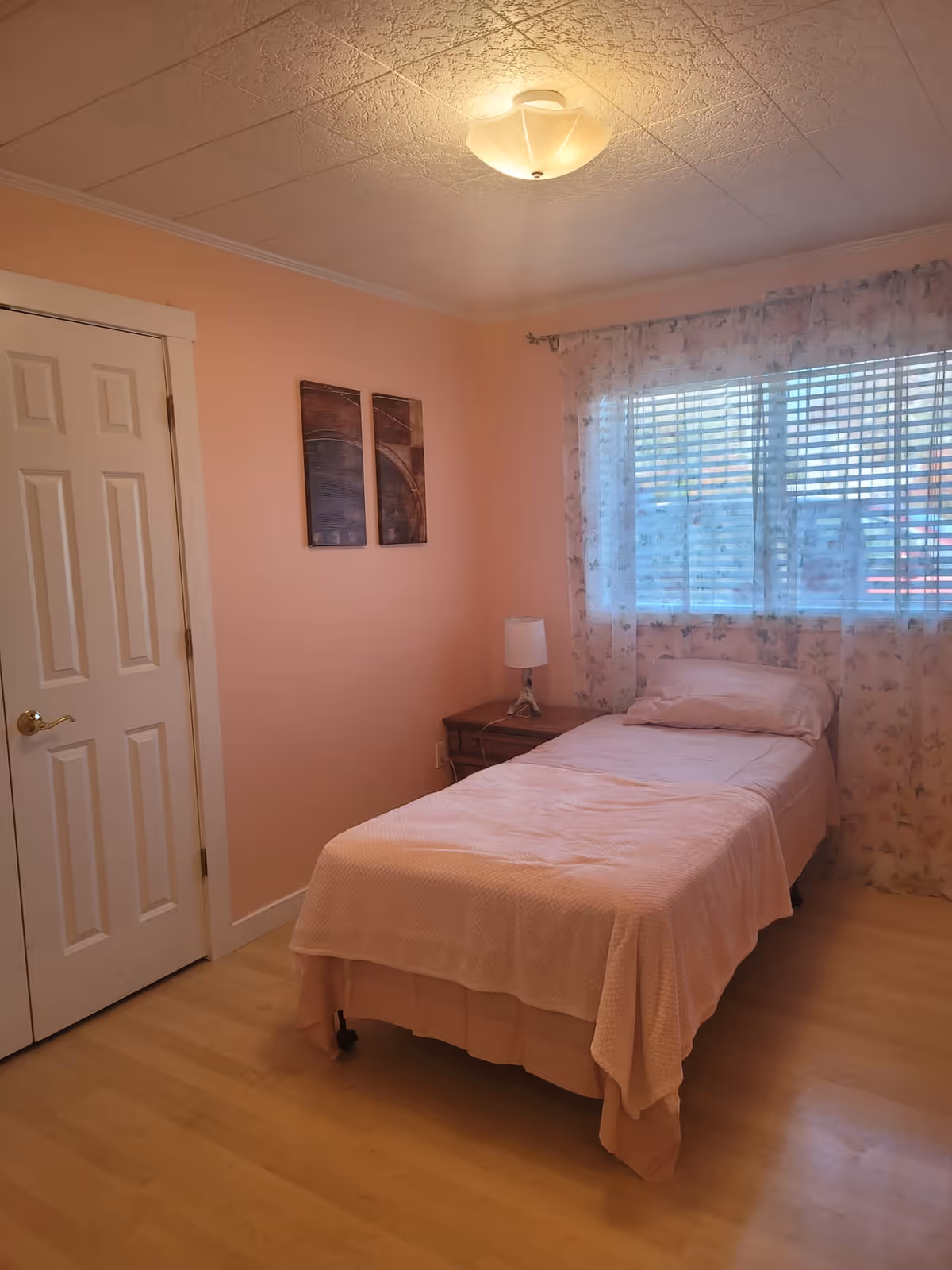 A small bedroom with peach-colored walls and light wood flooring. There is a single bed with a pink bedspread and pillow, positioned against a window with floral curtains. Next to the bed is a wooden nightstand with a white lamp. Two abstract paintings hang on the wall near a closed white door.