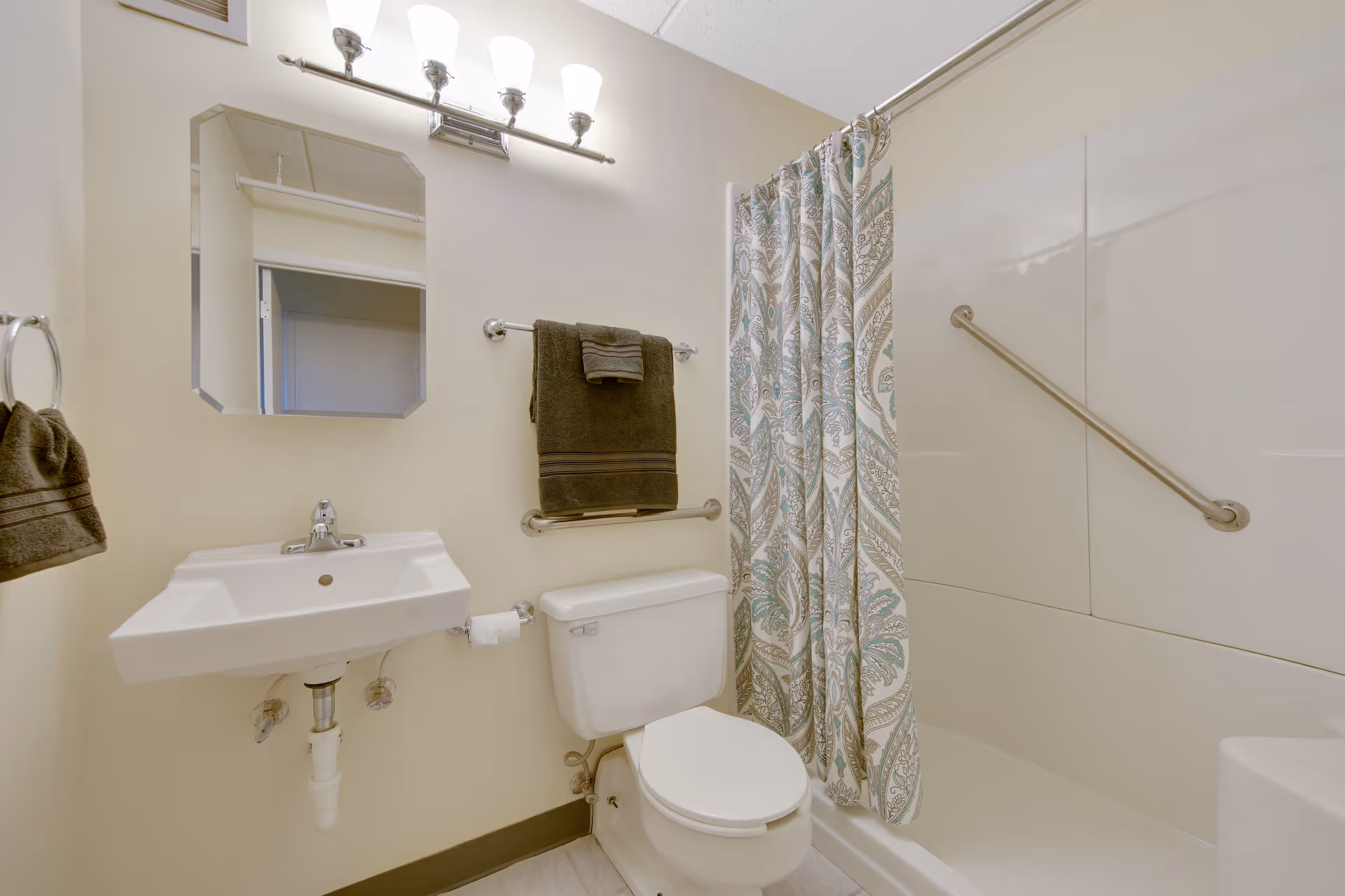 Accessible bathroom with a wall-mounted sink, toilet, towel racks, and a shower-tub with grab bars and a patterned curtain.