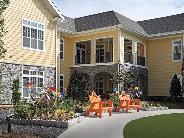 Outdoor view of a senior living facility with yellow siding and stone accents. There is a garden bed with colorful metal flower sculptures and plants. Two orange wooden chairs are placed on a curved concrete pathway near the garden. The building has a covered balcony with black railings and multiple windows.