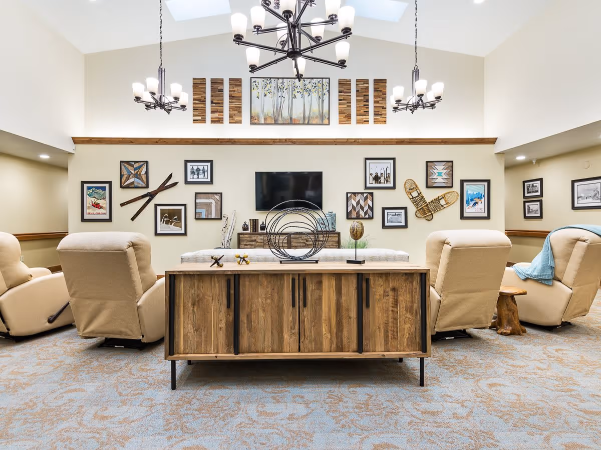 A cozy living room area in a memory care facility with beige recliner chairs arranged around a wooden cabinet. The wall features a mounted flat-screen TV surrounded by various framed pictures and decorative wooden art pieces, including vintage skis and snowshoes. The room has a high ceiling with skylights and modern chandeliers providing light.