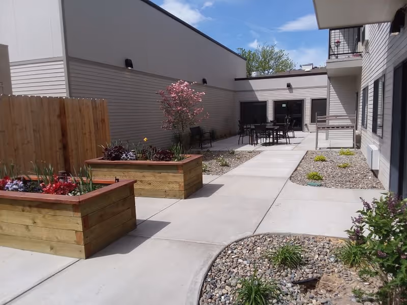 Courtyard with raised wooden planter boxes, paved walkways, patio tables and chairs, and surrounding building walls.