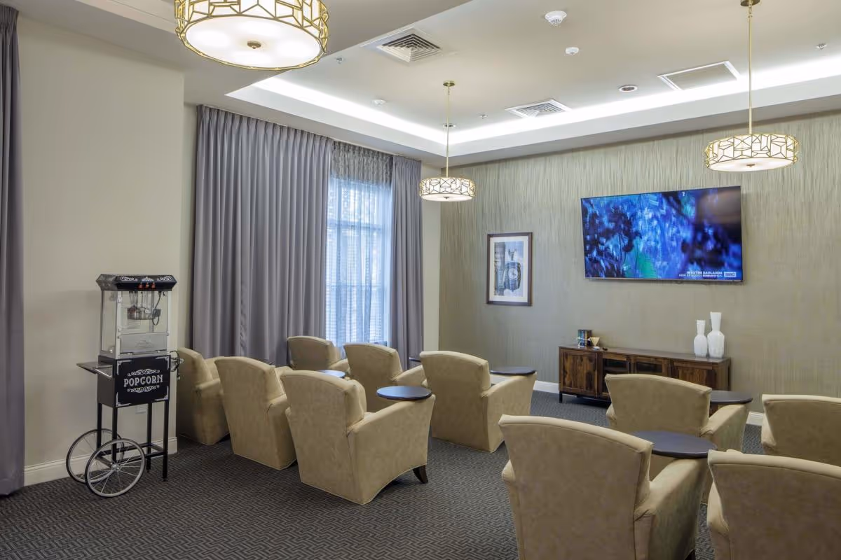 A cozy common area with beige armchairs arranged in rows facing a wall-mounted flat screen TV. The room has a popcorn machine on the left side, large windows with gray curtains, modern pendant lights hanging from the ceiling, and a wooden cabinet beneath the TV with decorative vases and framed artwork on the wall.