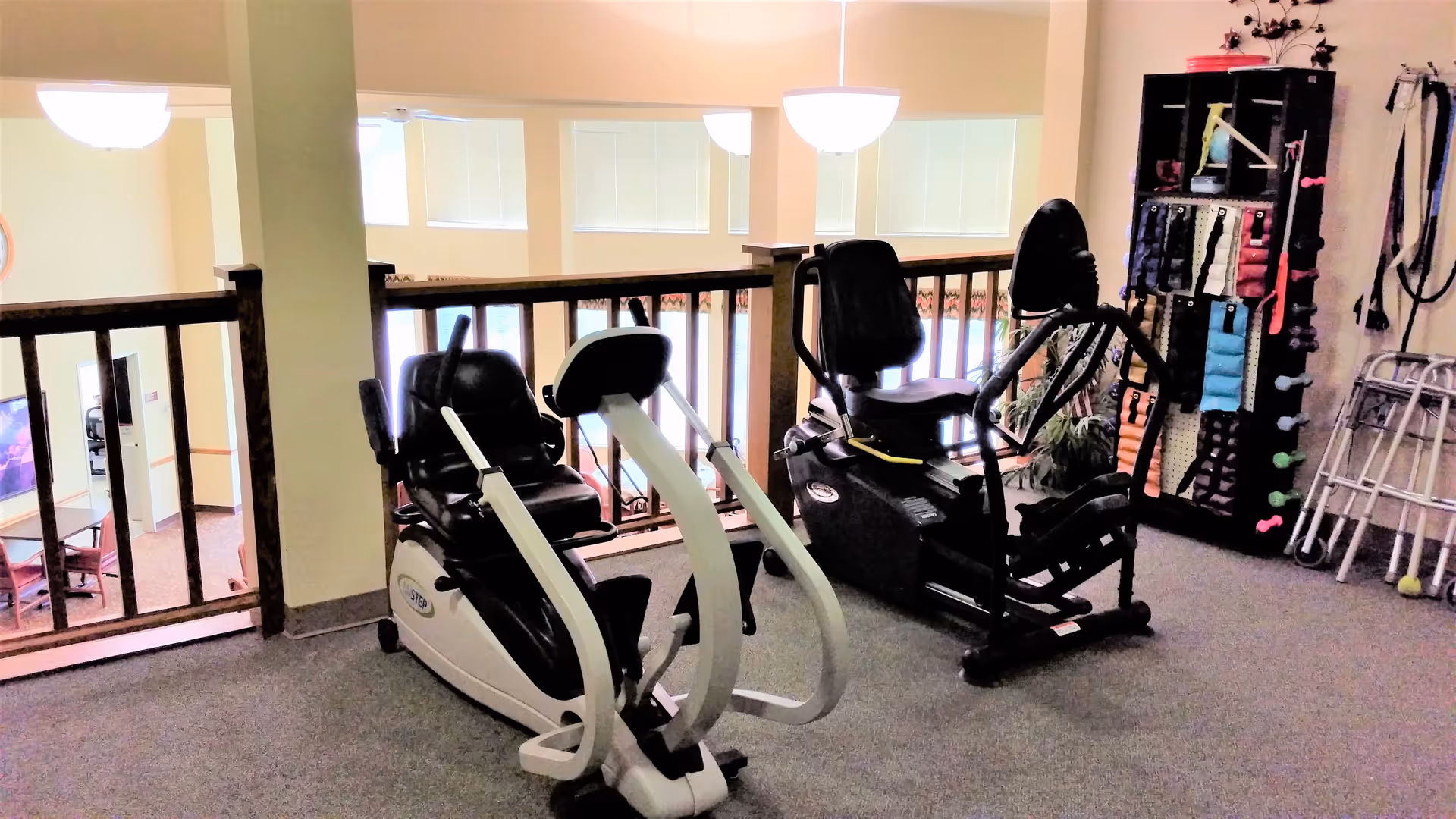 Two recumbent exercise bikes and a rack of small fitness equipment on a carpeted mezzanine in a senior living facility.