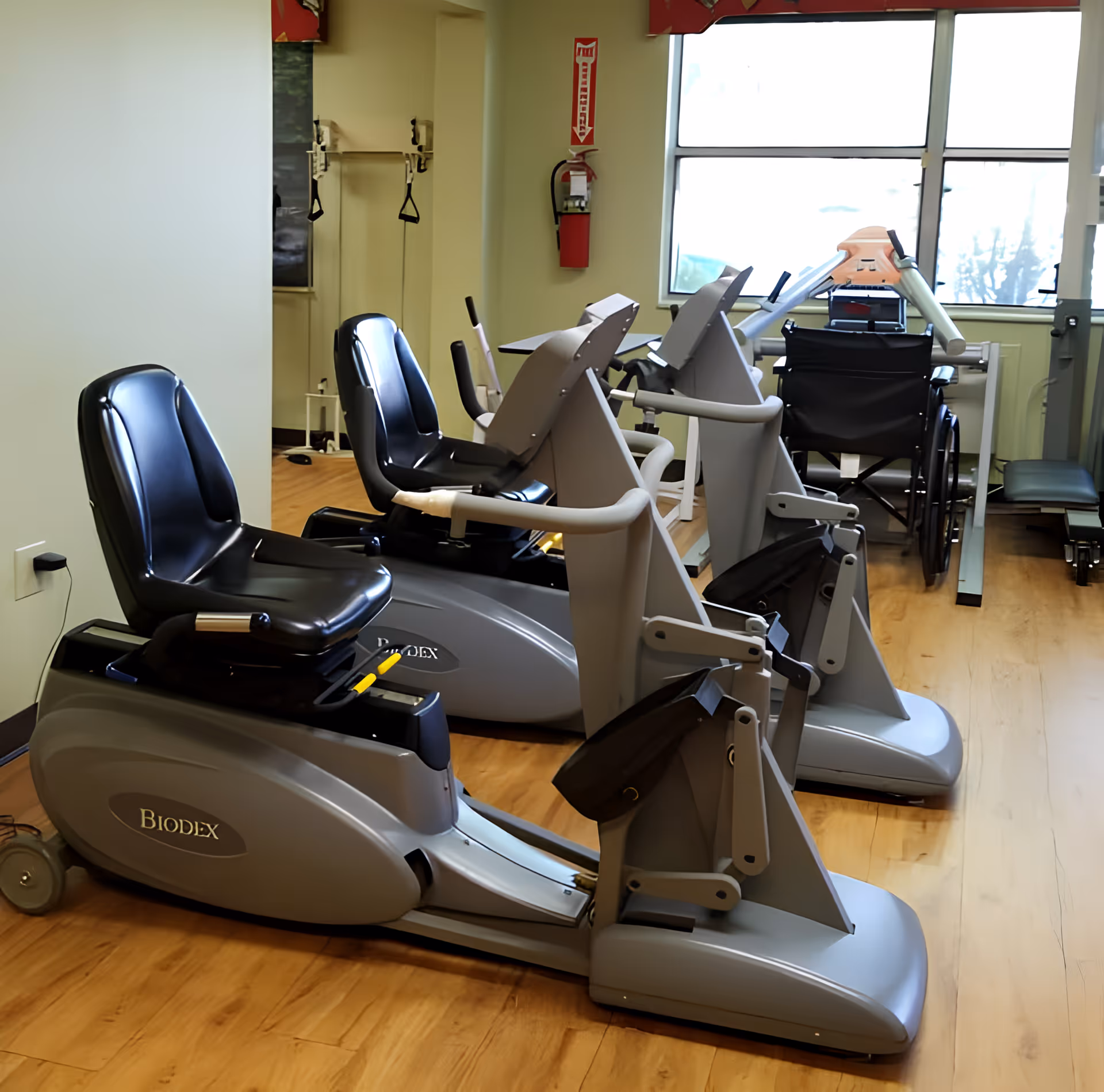 Rehabilitation gym room with multiple recumbent exercise machines and a wheelchair by a window.
