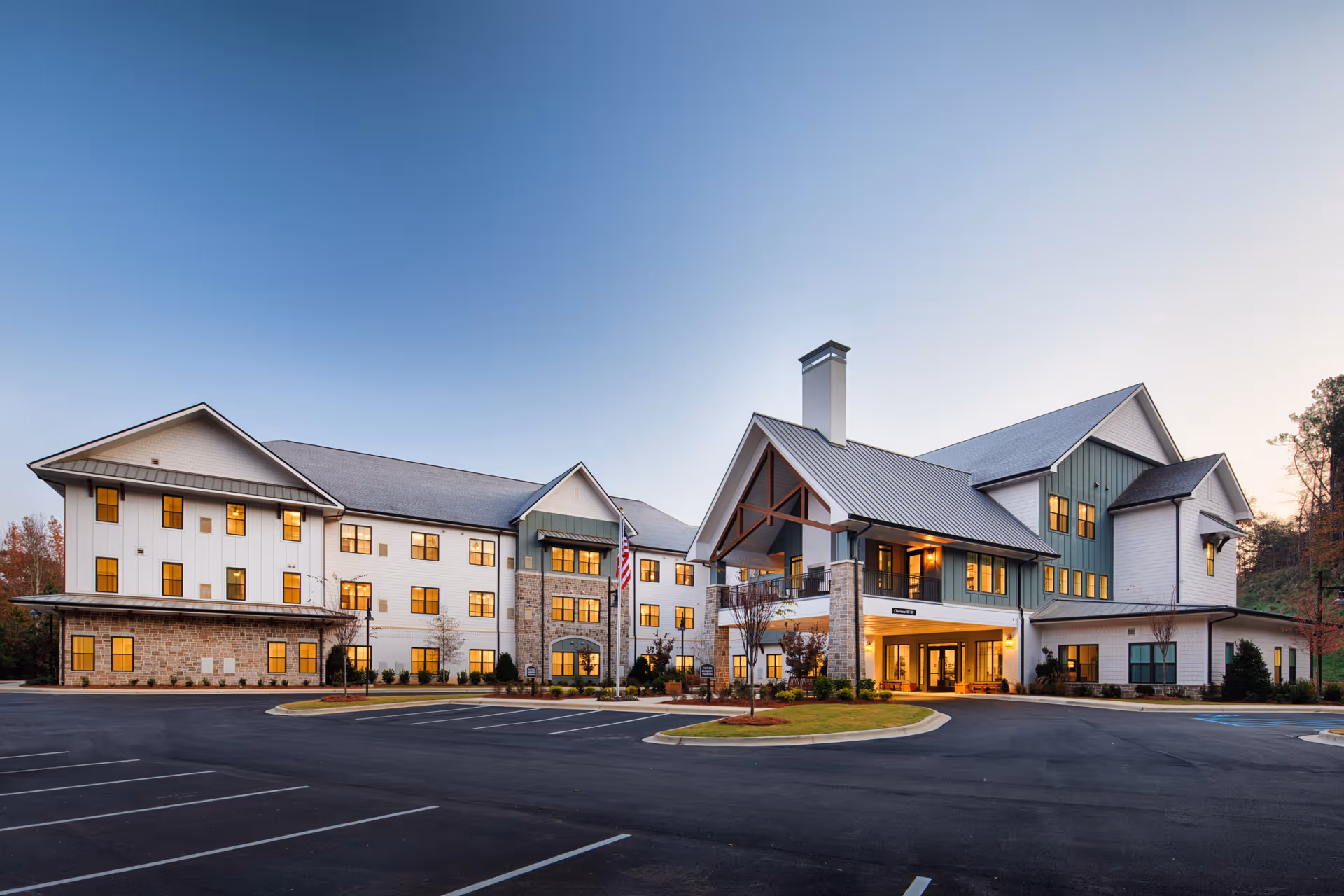 Exterior view of a large, modern senior living facility building named Longleaf Liberty Park during dusk, with warm lights glowing from the windows and a mostly empty parking lot in front.