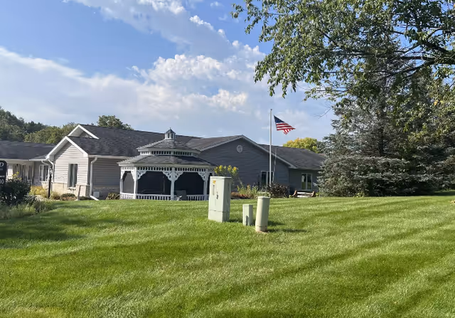 A single-story building with gray siding and a dark roof, featuring a white gazebo in front. There is a well-maintained green lawn with utility boxes and an American flag on a flagpole. Trees and blue sky with clouds are visible in the background.