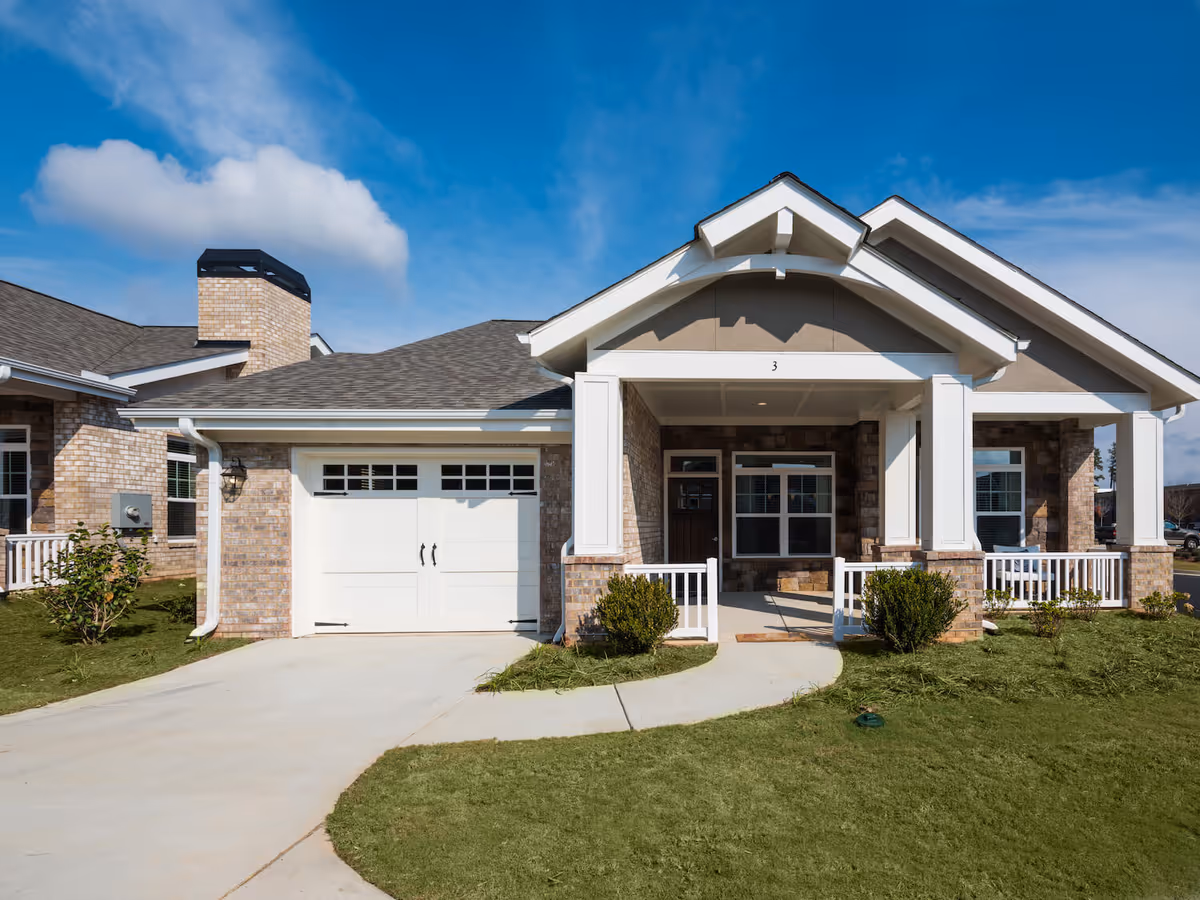 Exterior view of a single-story brick building with a white garage door, a covered front porch with white columns, and a well-maintained lawn under a blue sky with some clouds.