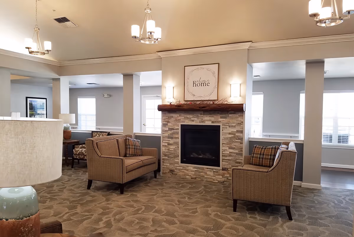 A cozy senior living facility common area with two beige armchairs facing a stone fireplace. Above the fireplace is a framed sign that reads 'welcome home'. The room has soft lighting from ceiling fixtures and wall sconces, with large windows letting in natural light.