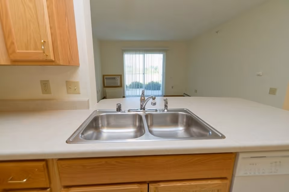 View of a kitchen countertop with a double stainless steel sink and a chrome faucet. Light wood cabinets are visible below and to the left of the countertop. In the background, there is a sliding glass door with vertical blinds and an air conditioning unit mounted on the wall.