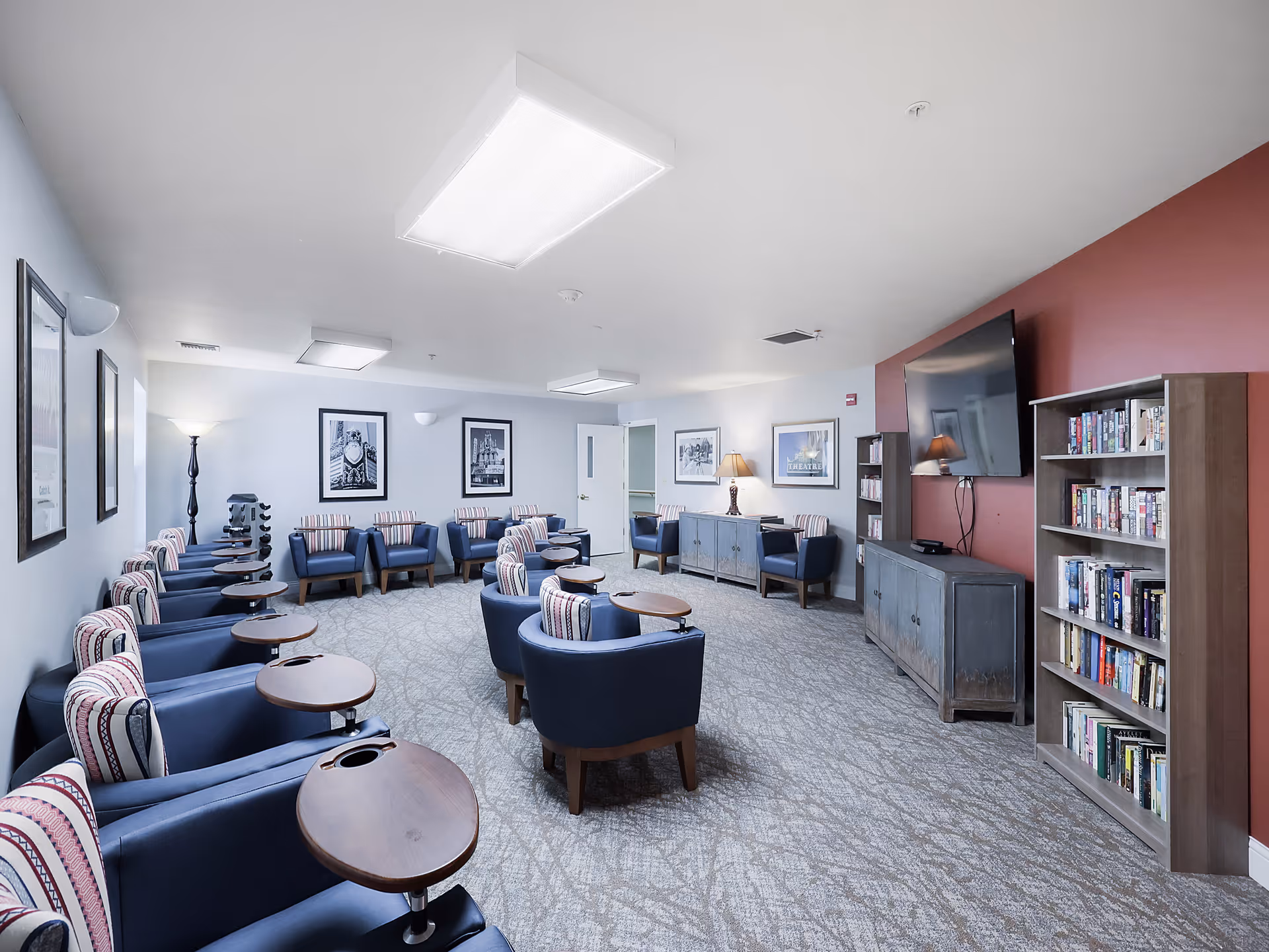 A spacious senior living common area with multiple blue armchairs arranged in rows, each with a small attached wooden table. The room has light gray walls with framed black and white pictures, a patterned carpet, a large flat-screen TV mounted on a red accent wall, bookshelves filled with books, and soft lighting from lamps and ceiling fixtures.