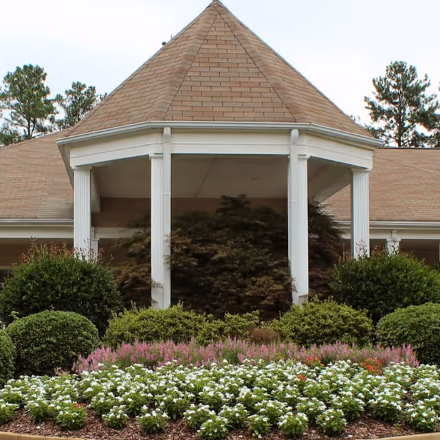 Exterior view of a building with a peaked roof supported by white columns, surrounded by well-maintained bushes and flower beds with white and pink flowers.