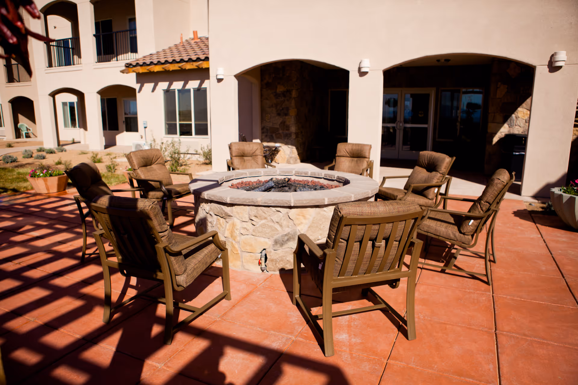 Outdoor patio area at The Rio Grande Gracious Retirement Living featuring a circular stone fire pit surrounded by eight cushioned chairs on a tiled floor with a building facade in the background.
