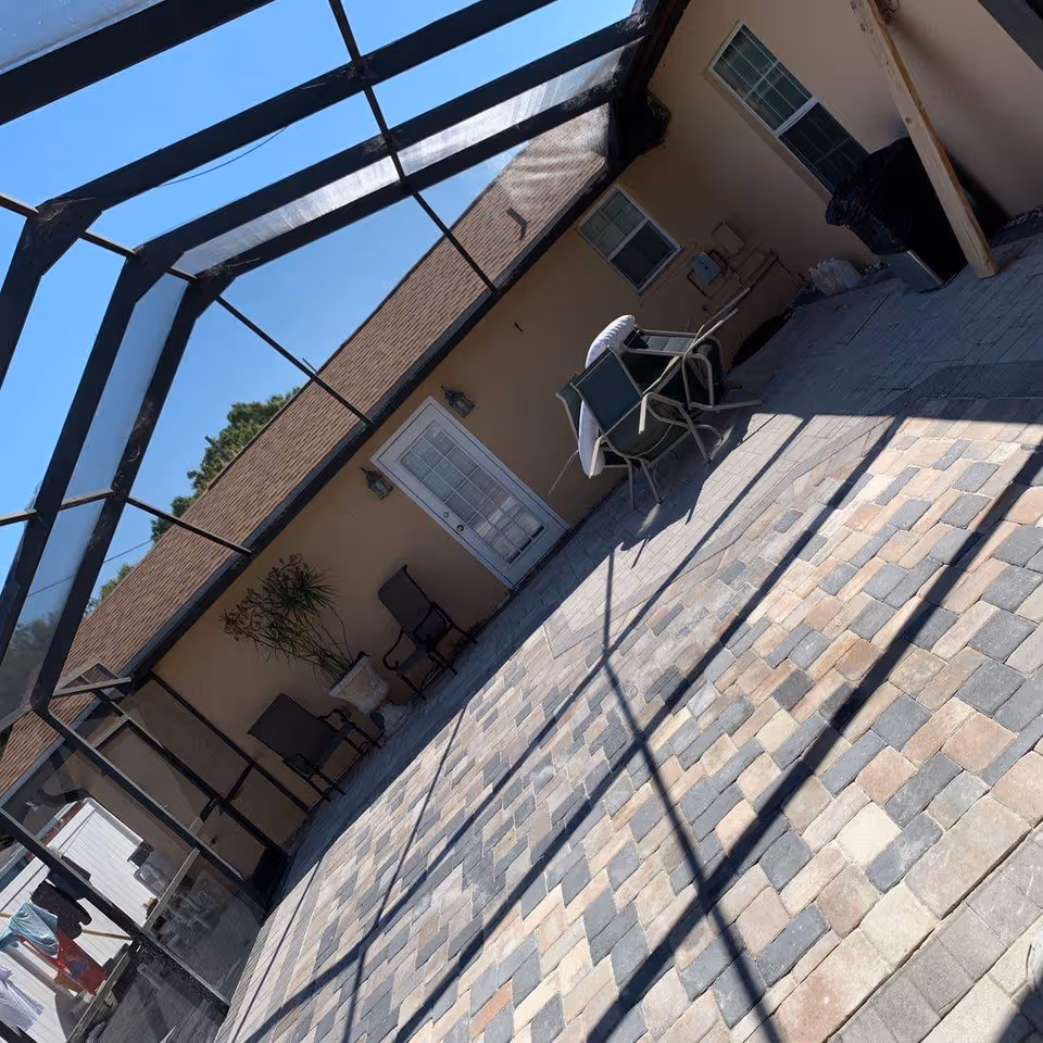 Outdoor patio area with a screened enclosure, paved stone floor, beige building wall with two windows and a glass door, several chairs, a potted plant, and some covered items.