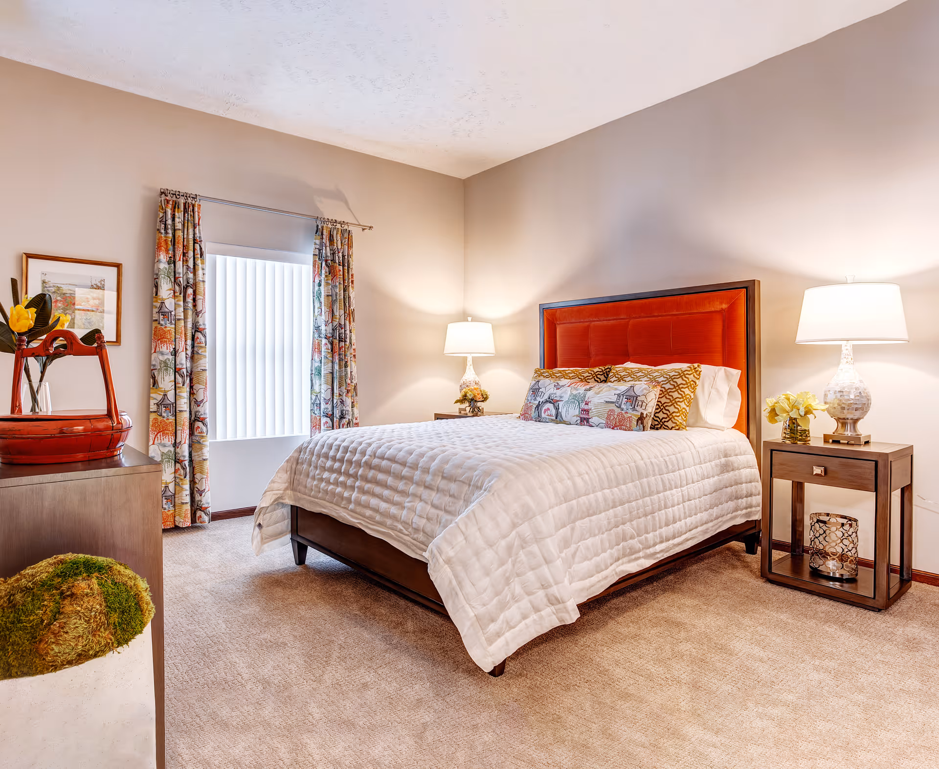 A cozy bedroom with a bed featuring a red upholstered headboard, white quilted bedding, and decorative pillows. There are two wooden nightstands on either side of the bed, each with a lamp and a small flower arrangement. A window with colorful patterned curtains and vertical blinds is on the left wall. A dresser with a red decorative item and a framed picture above it is also visible. The room has beige carpet and neutral-colored walls.